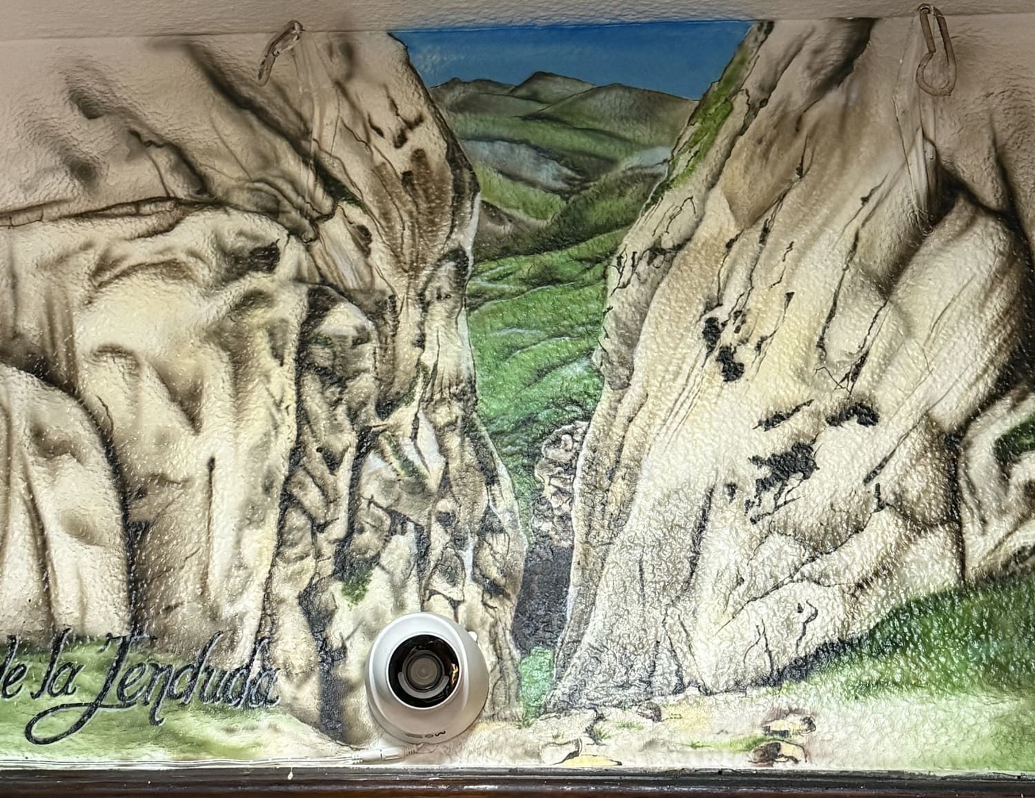 Mural que representa un cañón rocoso con un valle verde y cielo azul, con un desagüe circular en la parte inferior central.