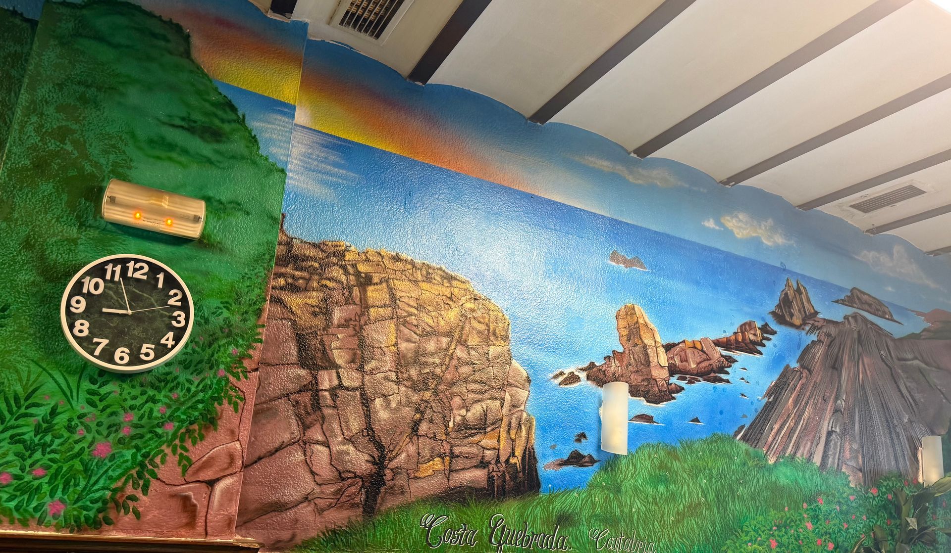 Mural colorido de montañas, cascada, pájaros y vegetación en una pared con un reloj a la izquierda.