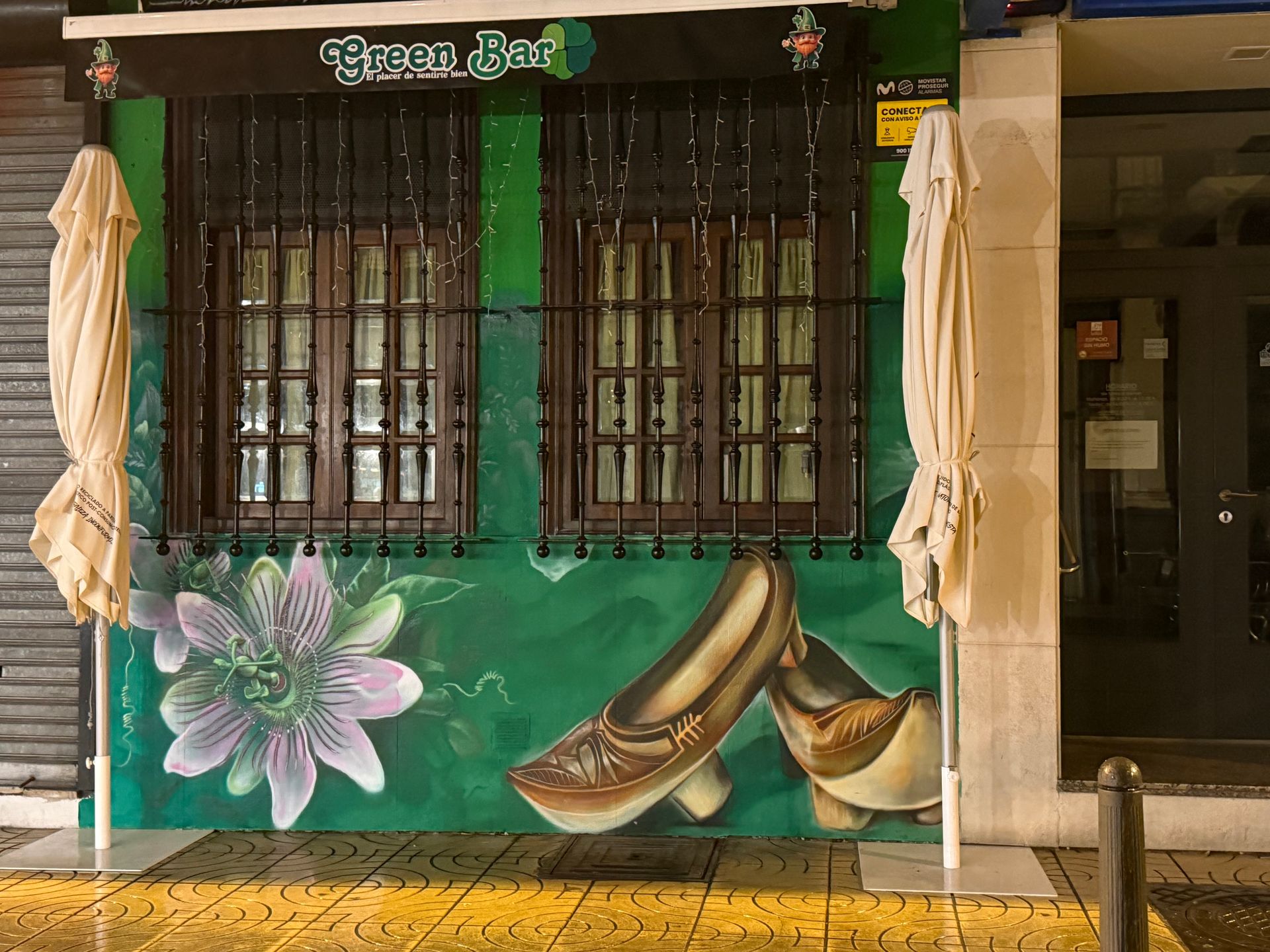 Fachada verde con murales pintados de flores y cuernos bajo ventanas enrejadas, flanqueada por cortinas beige enrolladas.