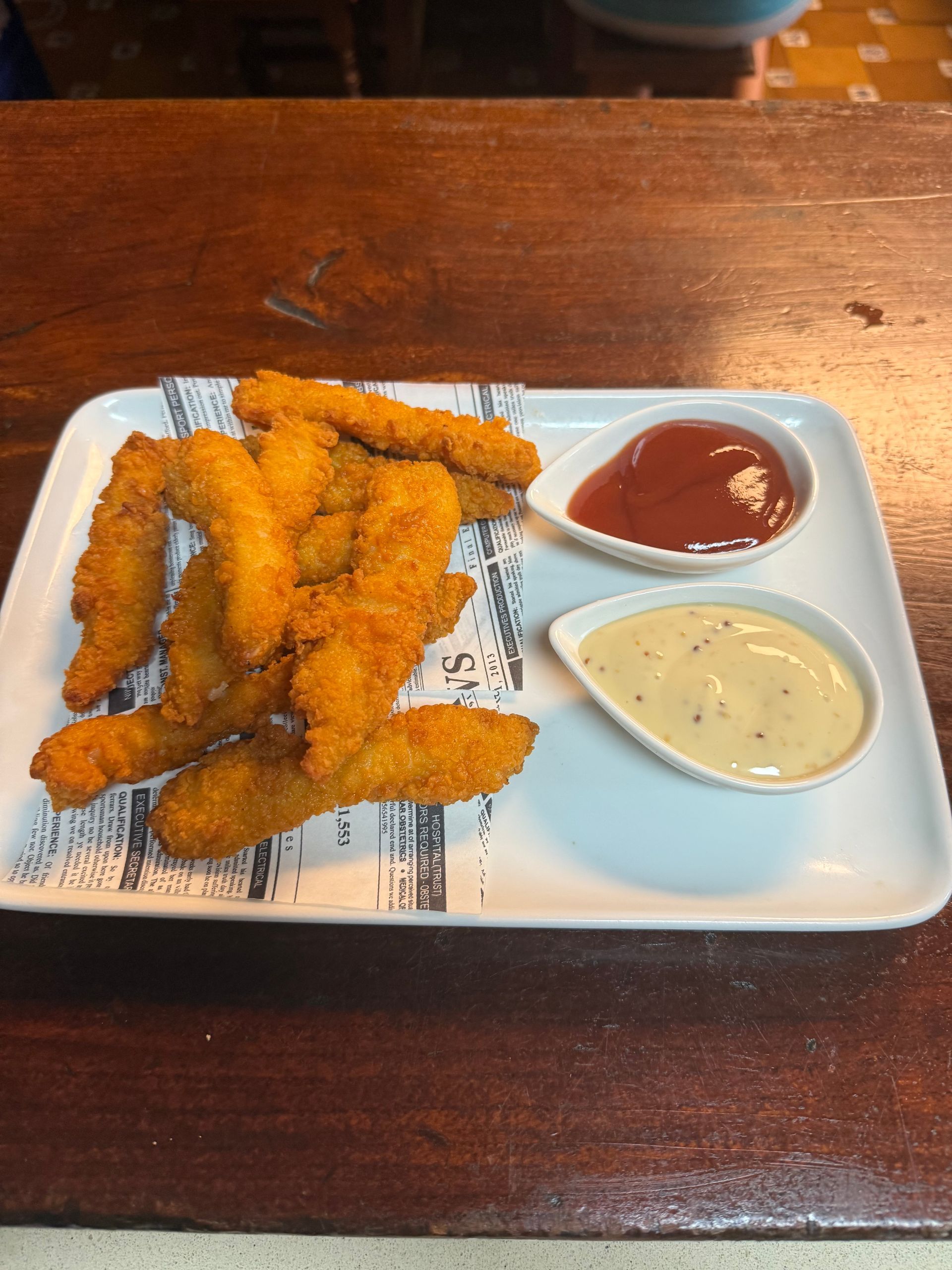Bandeja de tiras de pollo frito con ketchup y salsa para mojar en un plato blanco sobre una mesa de madera.