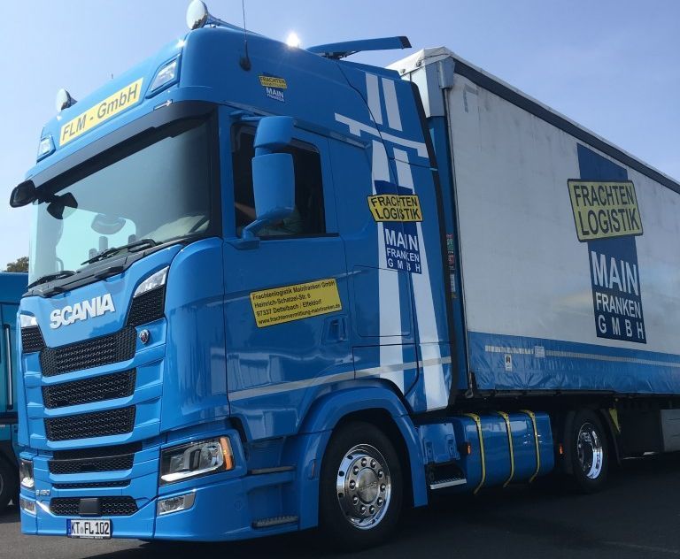 Blau-weißer MAN-LKW mit dem Logo der Frachten Logistik Main GmbH, geparkt an einem sonnigen Tag.