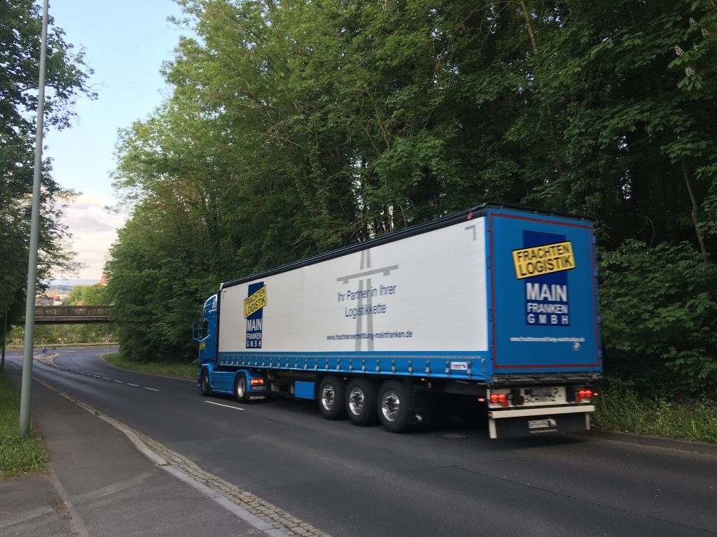 Blau-weißer Sattelschlepper fährt auf der Straße an Bäumen vorbei. Auf dem Anhänger steht „Main Transport“.