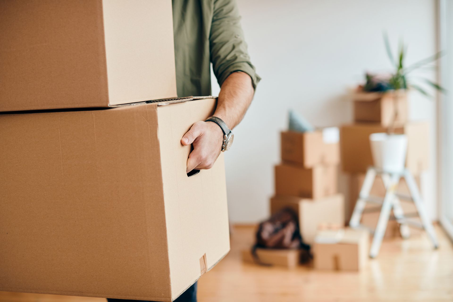 Una persona transporta cajas de cartón para mudanza en una habitación luminosa con más cajas y una silla al fondo.