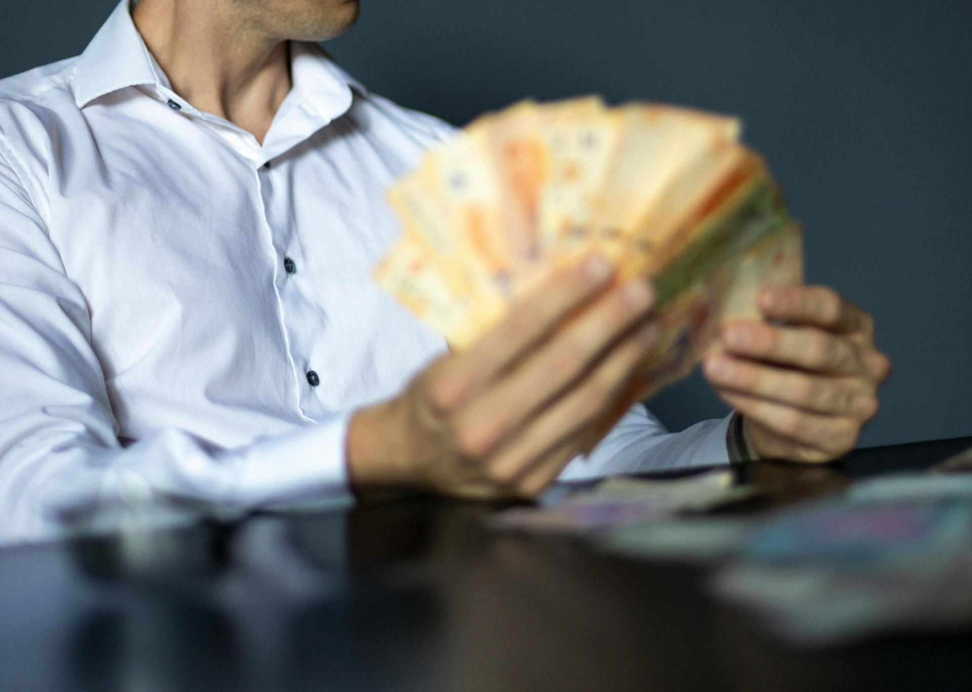Una persona con una camisa blanca abotonada sostiene y extiende un fajo de billetes sobre una mesa oscura.