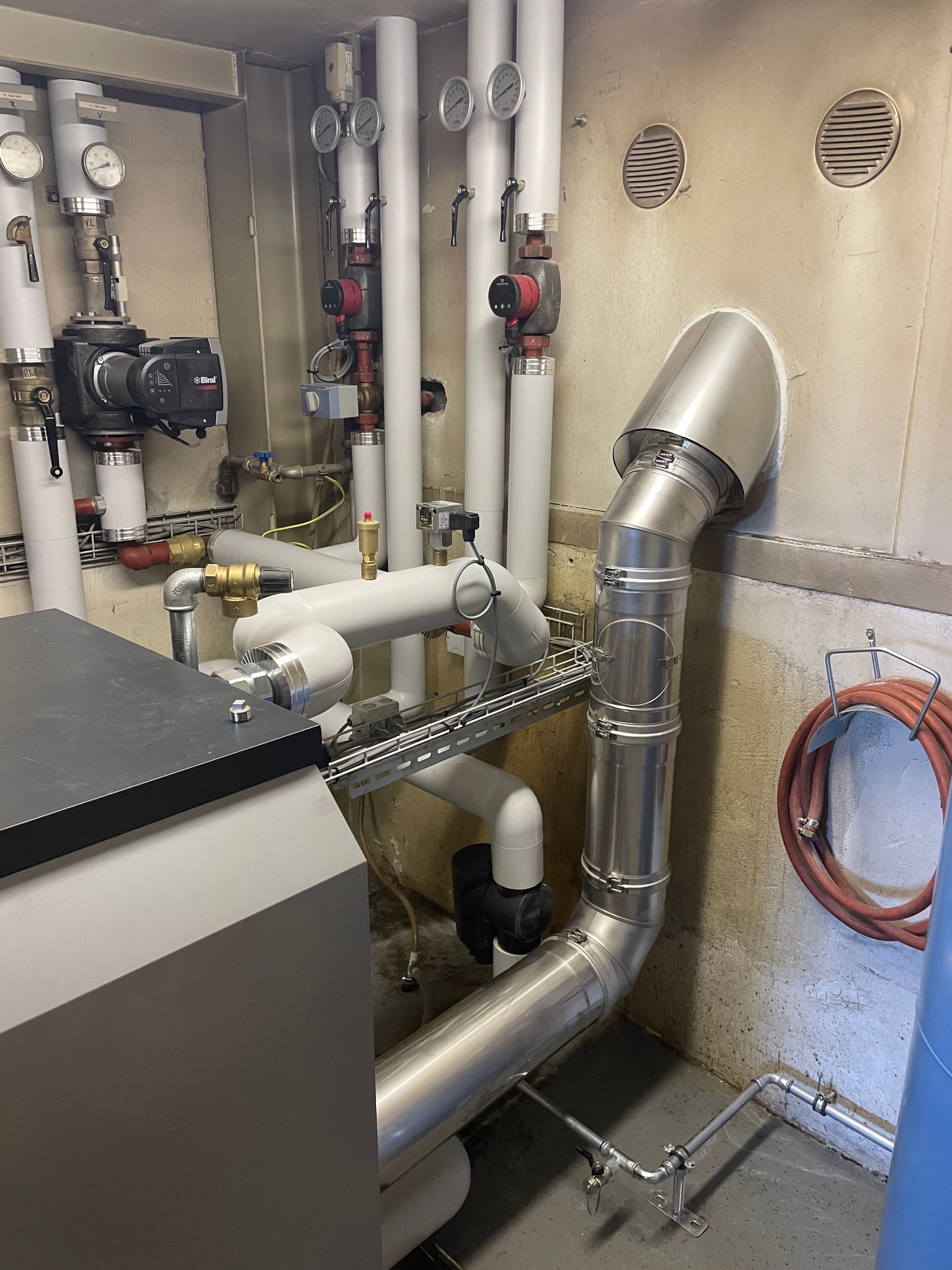 Person mit Handschuhen wartet eine Ausseneinheit einer Klimaanlage mit Manometern; Hintergrund ist eine rosa Wand zu sehen.