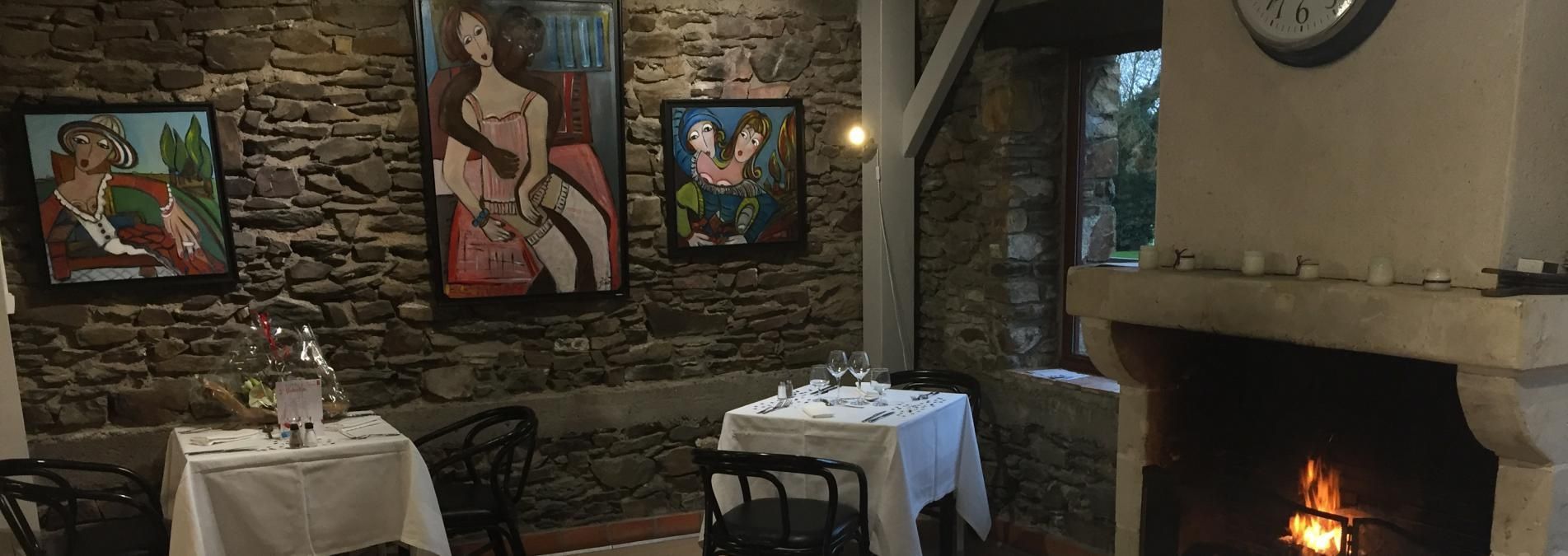 Acogedor comedor con paredes de piedra, cuadros enmarcados, mesas con manteles blancos y una chimenea encendida a la derecha.