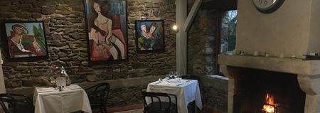 Salle à manger chaleureuse aux murs de pierre, agrémentée de tableaux encadrés, d'une petite table dressée et d'une cheminée allumée sur la droite.