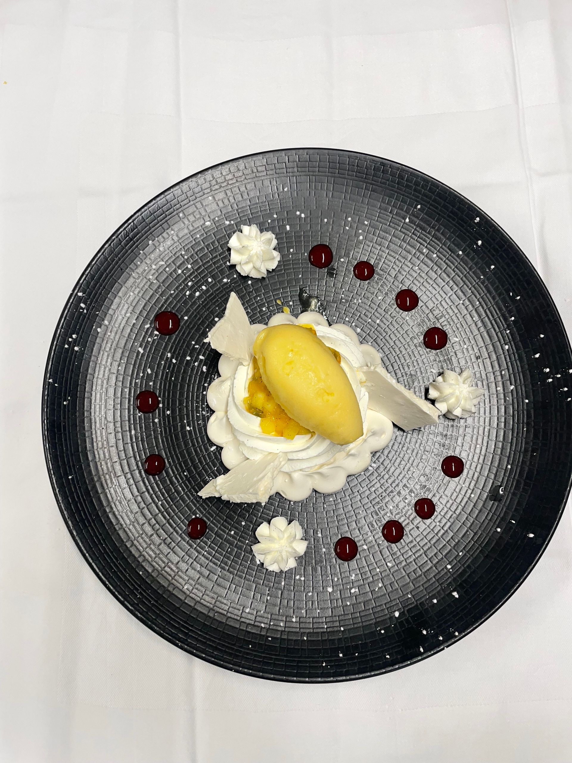 Assiette à dessert avec crème fouettée, sorbet jaune et points de coulis de fruits rouges sur une assiette ronde foncée