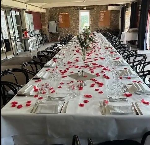 Une longue table de banquet dressée avec une nappe blanche, de la verrerie, des chaises et des pétales de roses rouges dans une pièce lumineuse