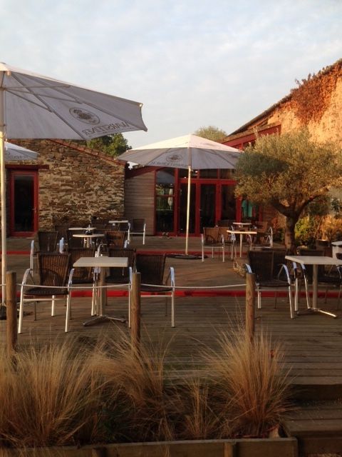 Terrasse extérieure de café avec tables, chaises, parasols et graminées ornementales à côté de bâtiments en pierre au coucher du soleil