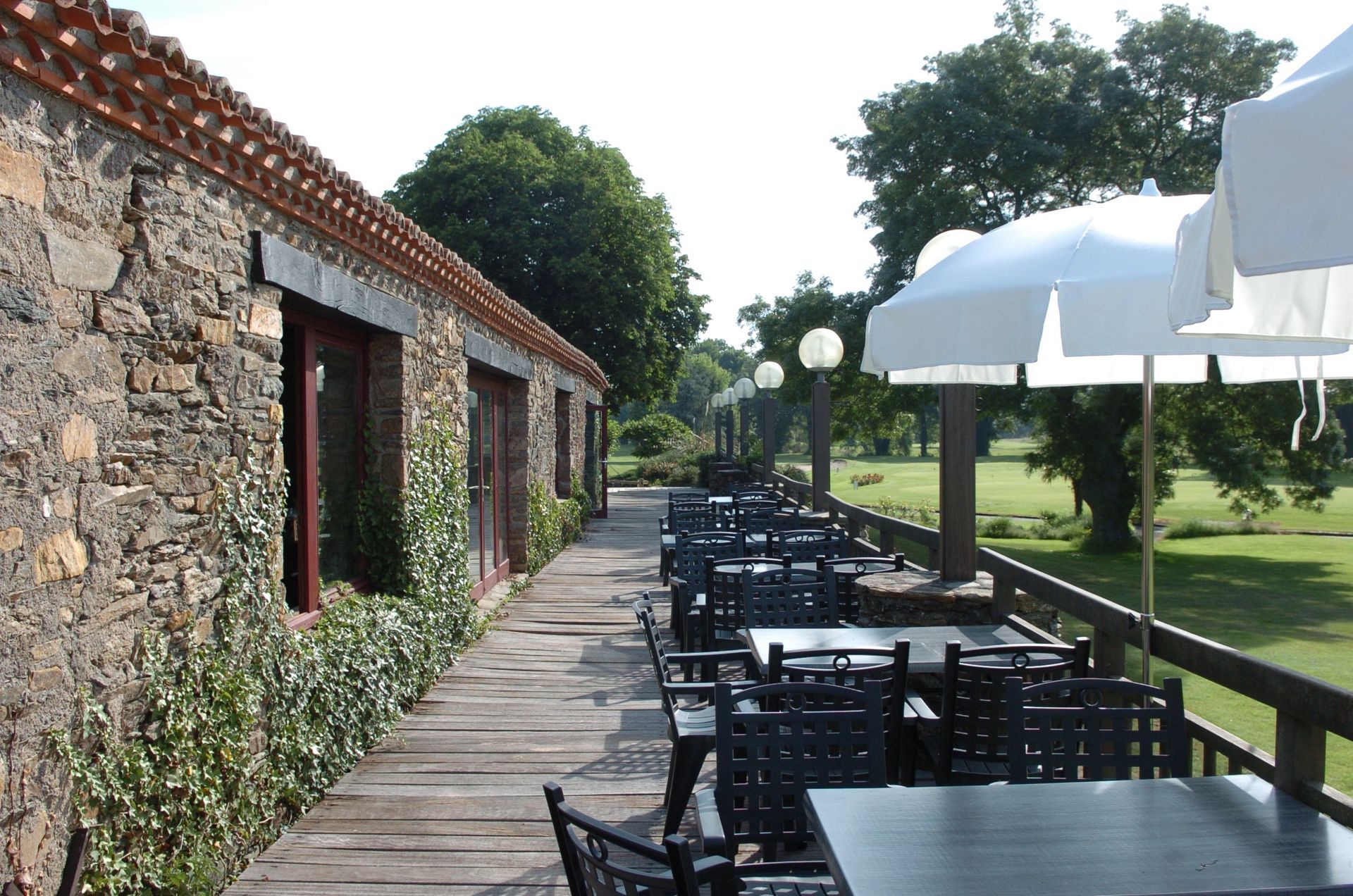 Terrasse extérieure attenante à un bâtiment en pierre, avec tables et chaises noires, et parasols blancs, le long d'une terrasse en bois.