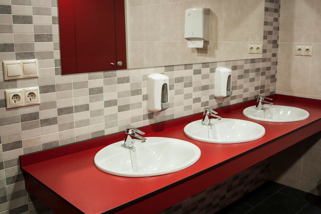 Baño público con tres lavabos, encimera roja, pared de azulejos grises, espejo y dispensadores de jabón.