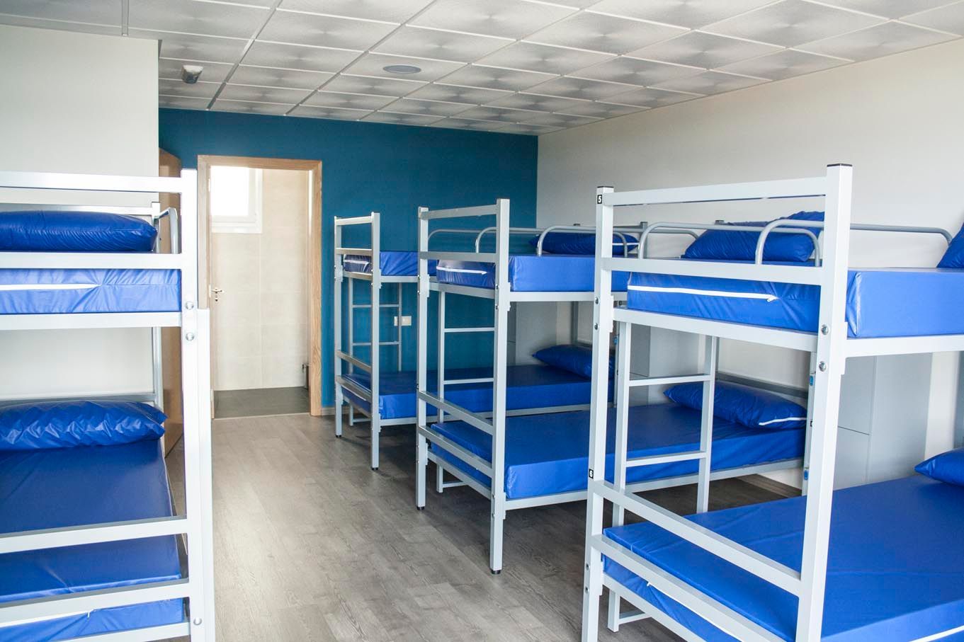 Literas en una habitación de hostal. Colchones azules, estructura metálica blanca, suelo gris claro y paredes azules