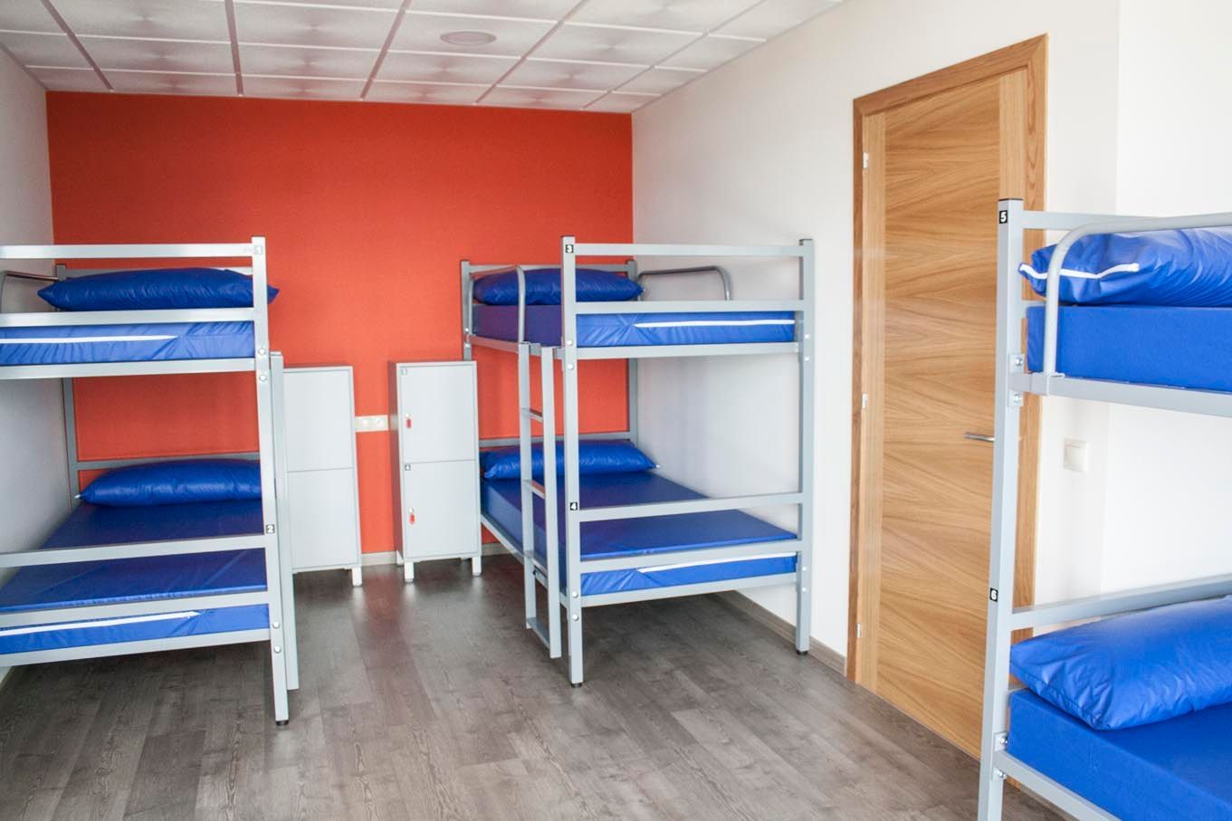 Literas en una habitación bien iluminada con colchones azules. Pared con acento rojo, taquillas y puerta de madera.