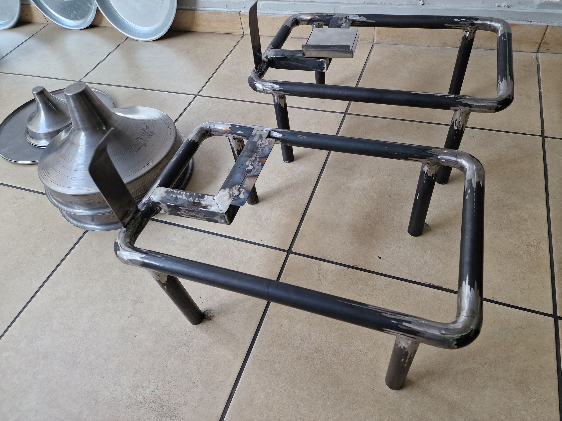 Δύο μαύρα μεταλλικά τραπεζομάντιλα, με πιάτα και μεταλλικά κωνικά αντικείμενα στο φόντο, τοποθετημένα σε πλακάκια δαπέδου.