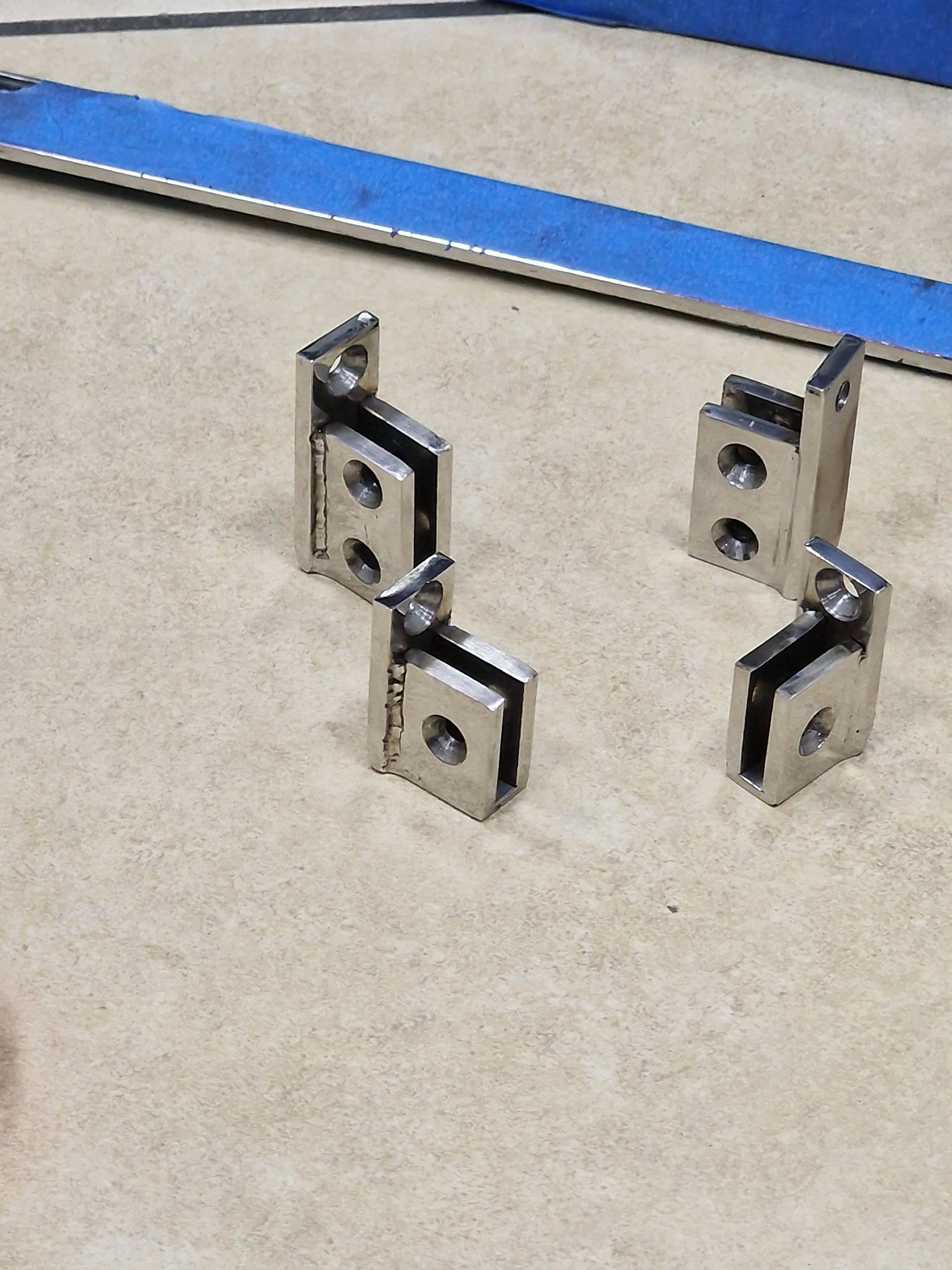 Τέσσερις ασημένιοι μεταλλικοί σφιγκτήρες γυαλιού σε μπεζ επιφάνεια, με μια μπλε λωρίδα στο φόντο.