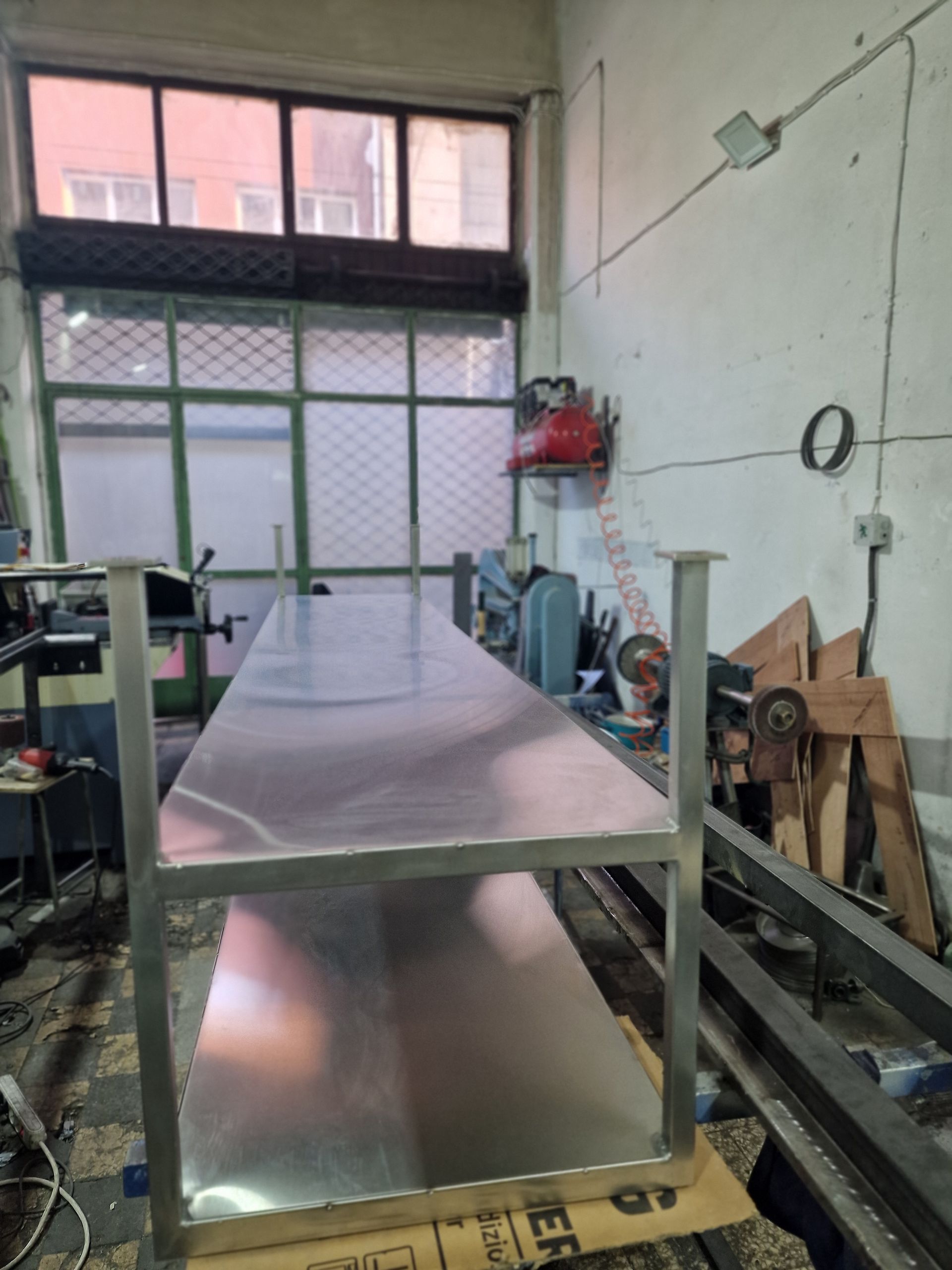 Πάγκος εργασίας από ανοξείδωτο ατσάλι σε εργαστήριο, ορατά πόδια, μακριά επιφάνεια τραπεζιού, με εργαλεία και ένα παράθυρο στο βάθος.