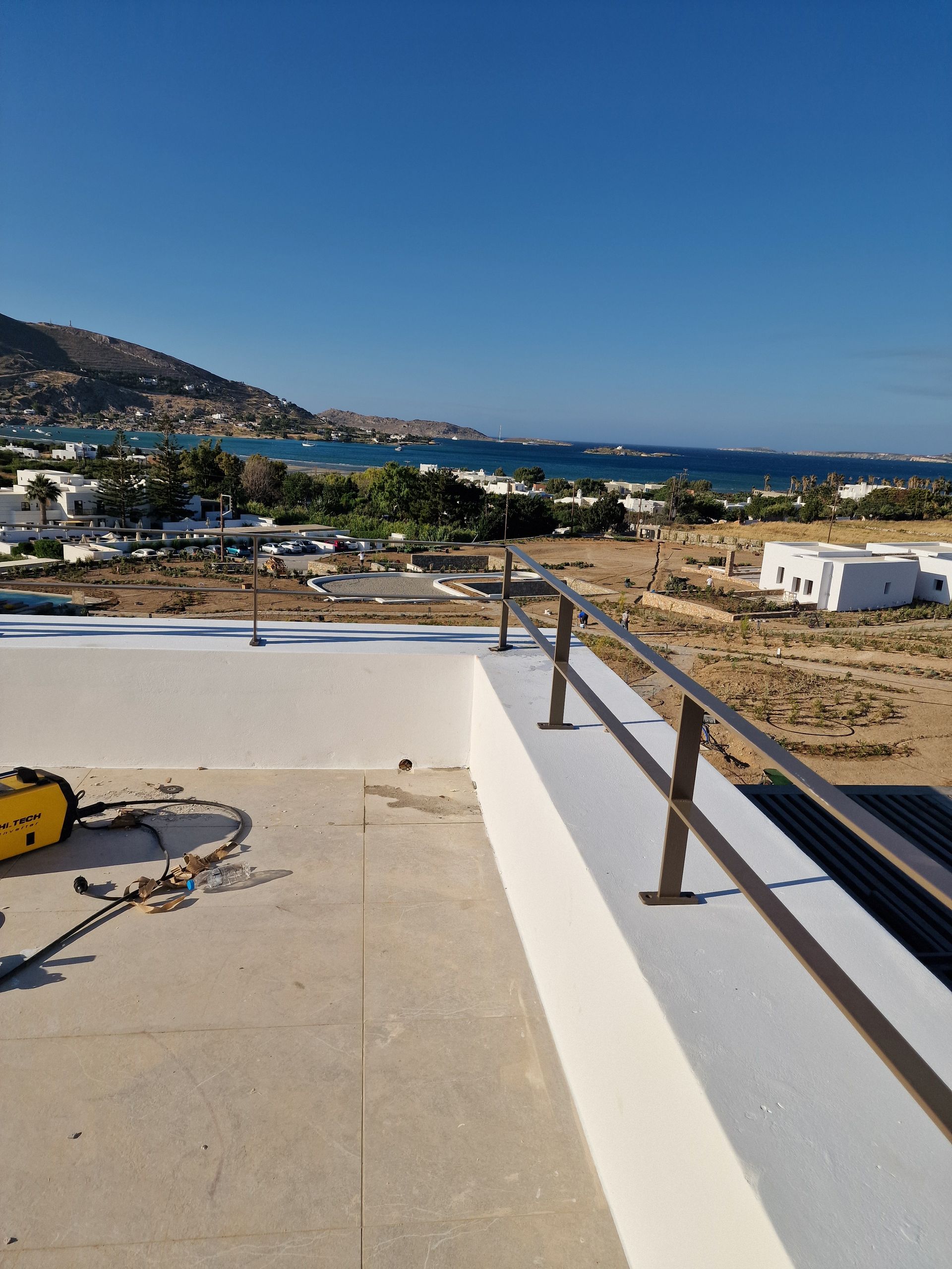 Ταράτσα με μεταλλικό κιγκλίδωμα με θέα σε ένα παράκτιο τοπίο κάτω από έναν γαλάζιο ουρανό.