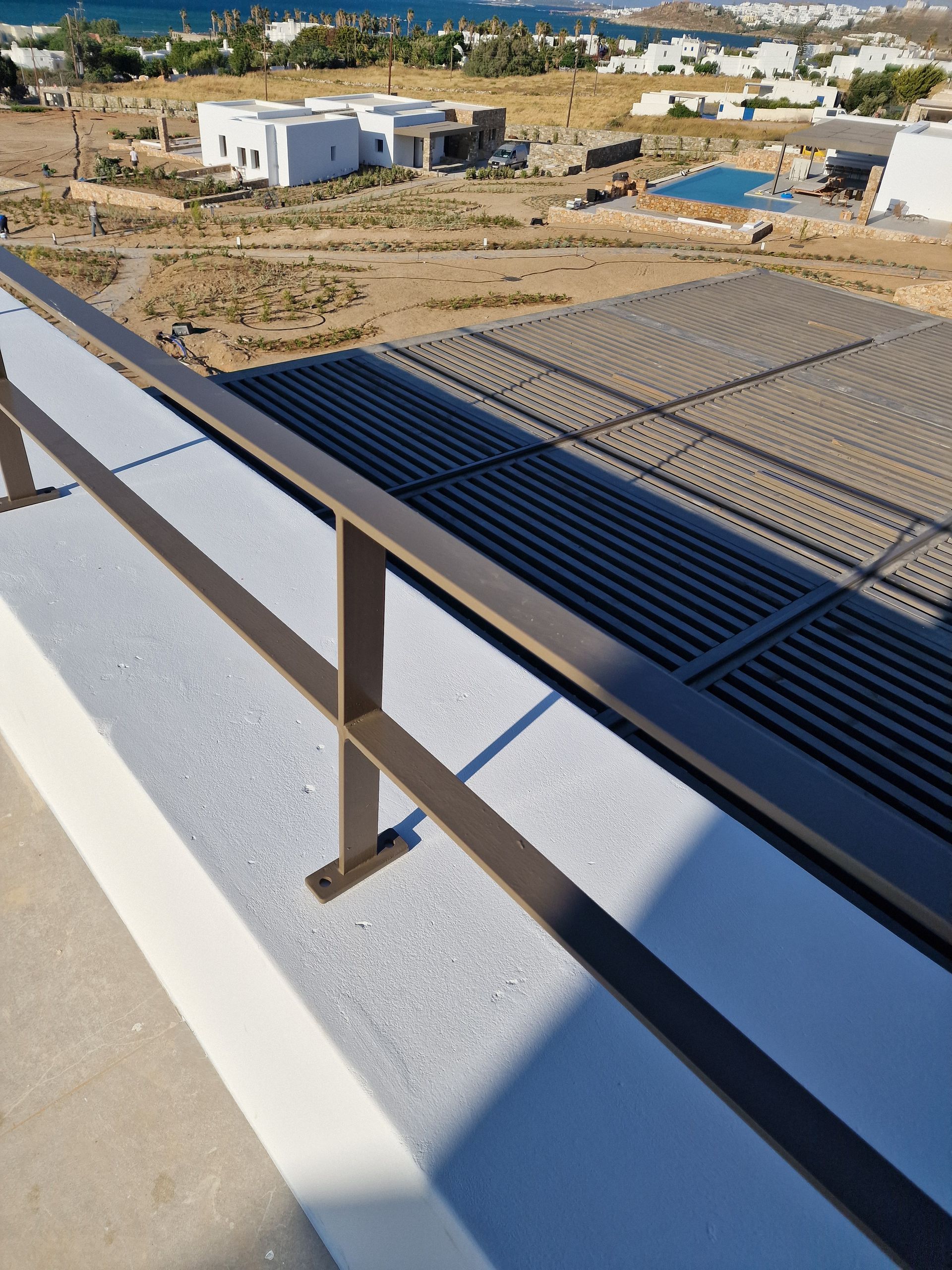 Καφέ κιγκλίδωμα σε λευκή επιφάνεια με μια επιφάνεια με πλέγμα από πίσω. Η θάλασσα και τα κτίρια βρίσκονται στο βάθος.