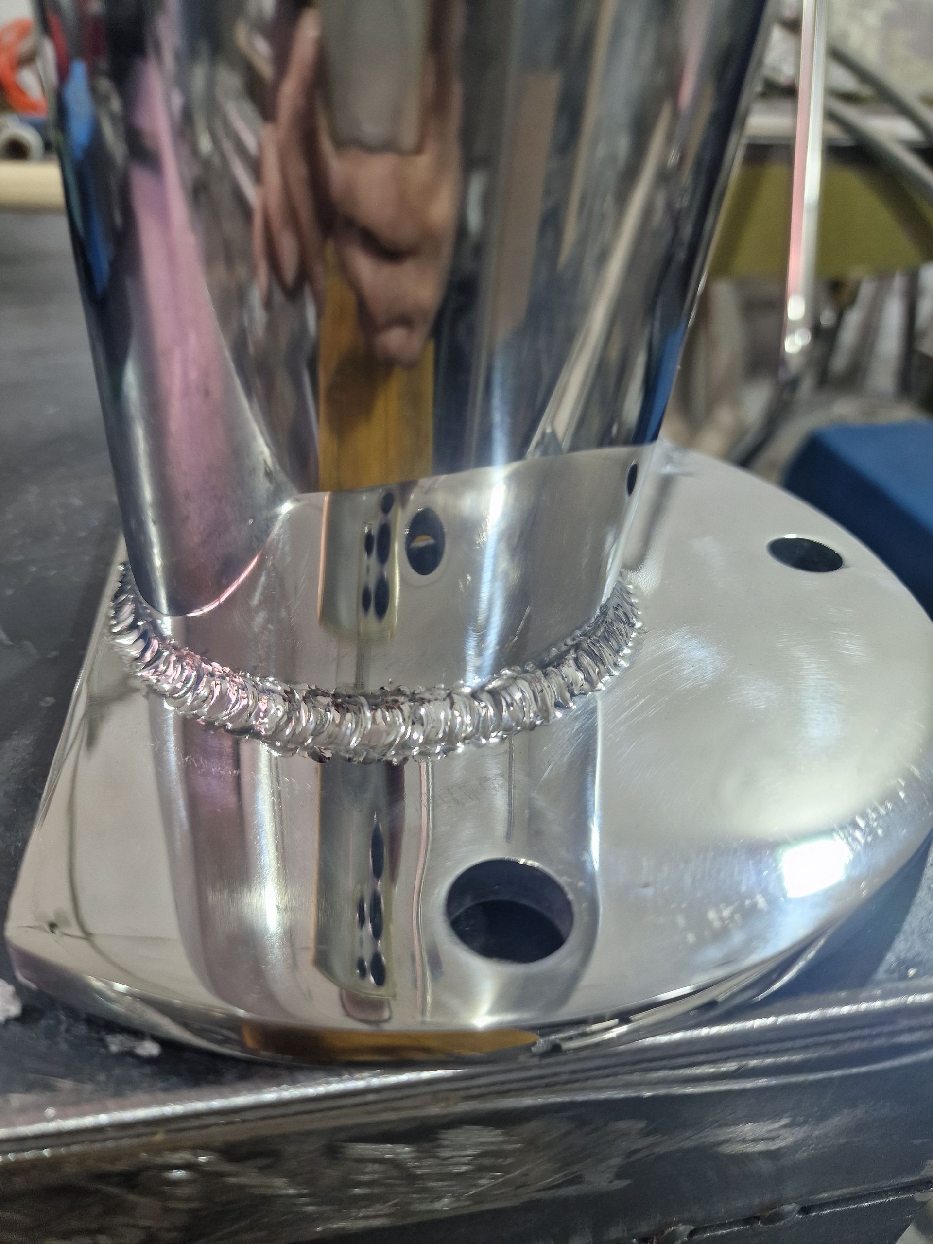 Κοντινό πλάνο ενός γυαλιστερού μεταλλικού αντικειμένου με συγκολλημένη ραφή, που δείχνει κυκλικές τρύπες και ανακλαστική επιφάνεια.