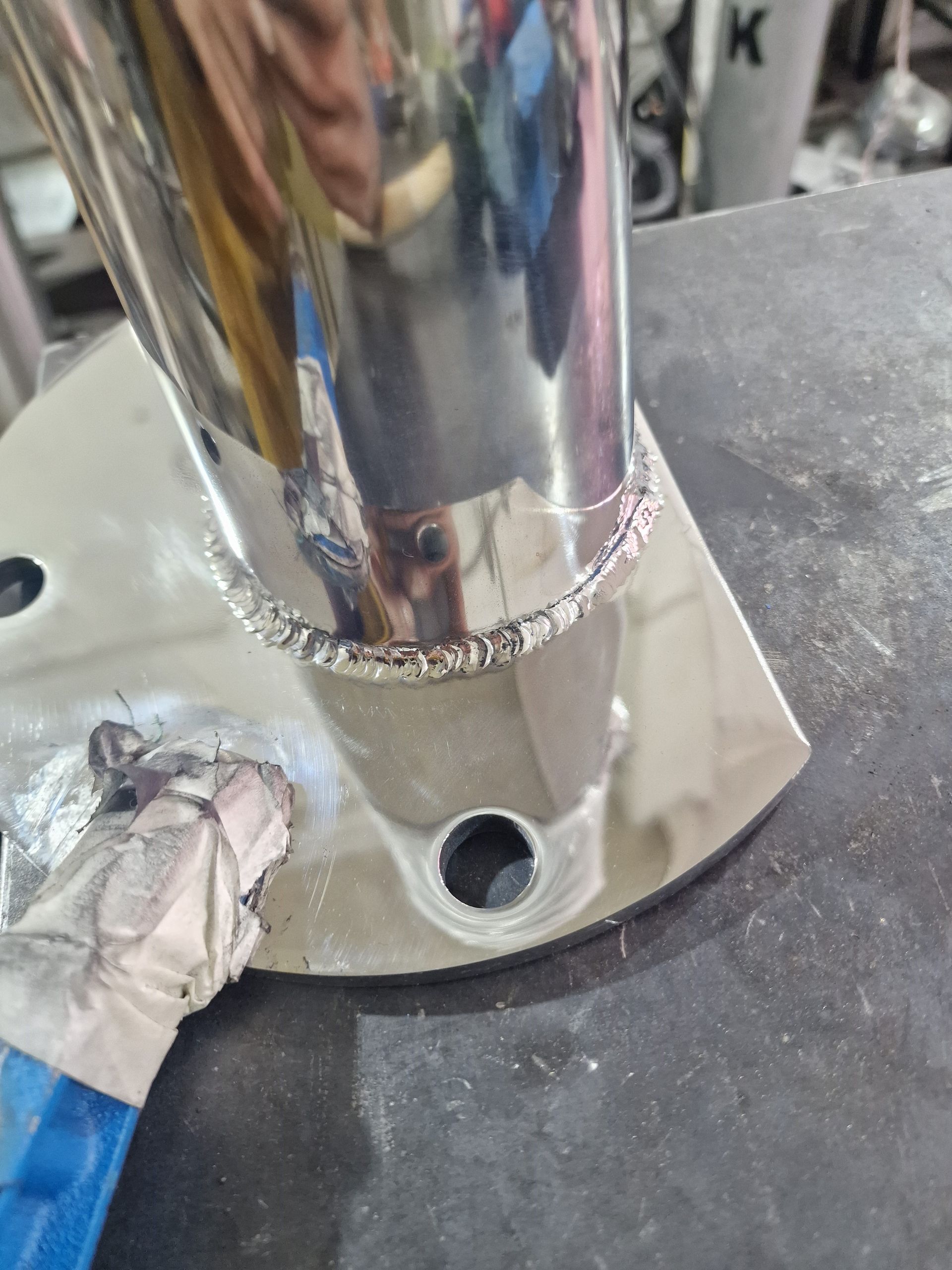 Συγκολλημένος μεταλλικός σωλήνας σε γυαλιστερή, επίπεδη μεταλλική βάση με τρύπες για μπουλόνια. Το γάντι ενός συγκολλητή κρατάει ένα εργαλείο.