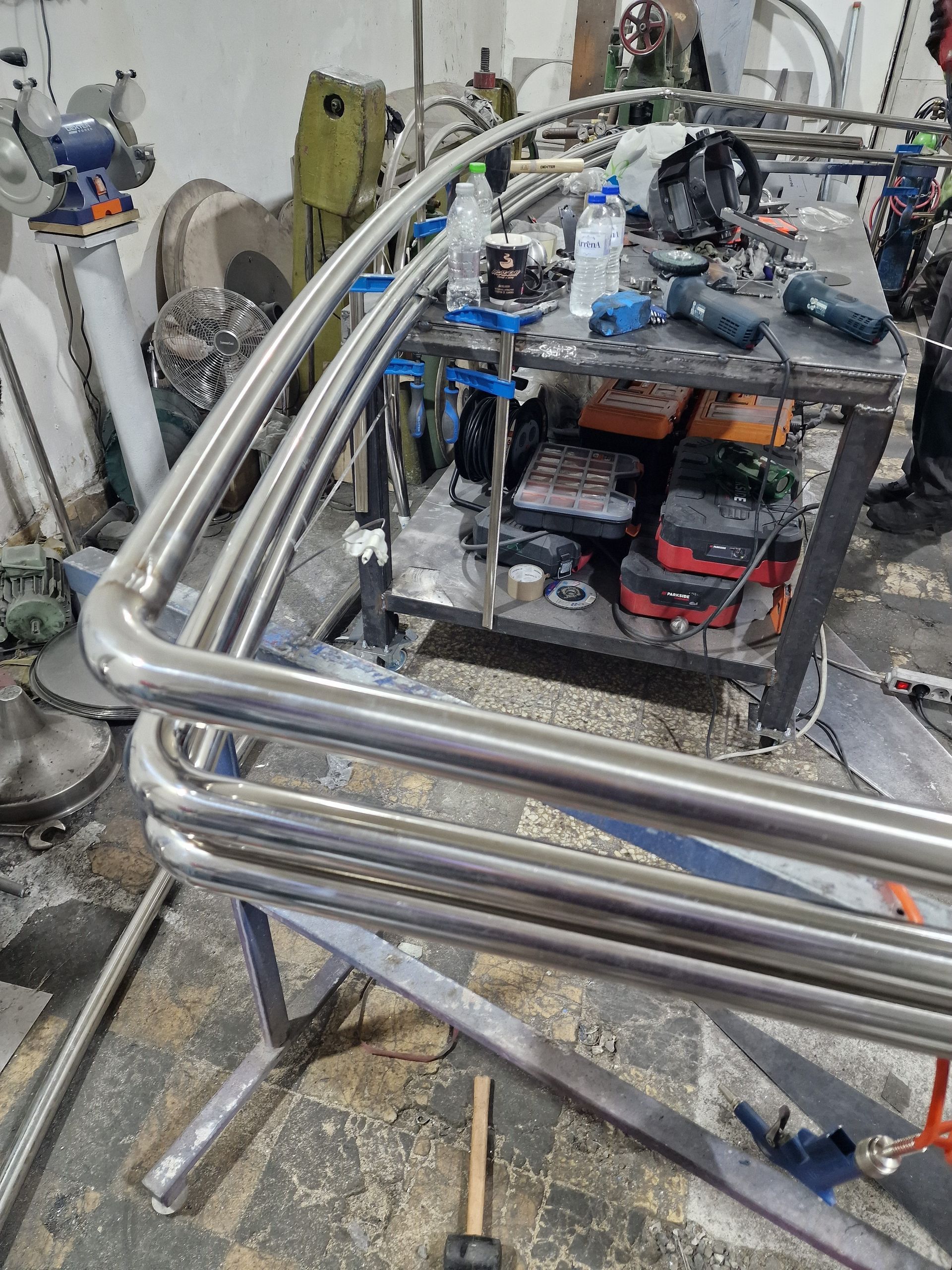 Μεταλλικός σκελετός σωληνώσεων υπό κατασκευή σε εργαστήριο· εργαλεία και εξοπλισμός ορατά σε ένα τραπέζι και στο φόντο.