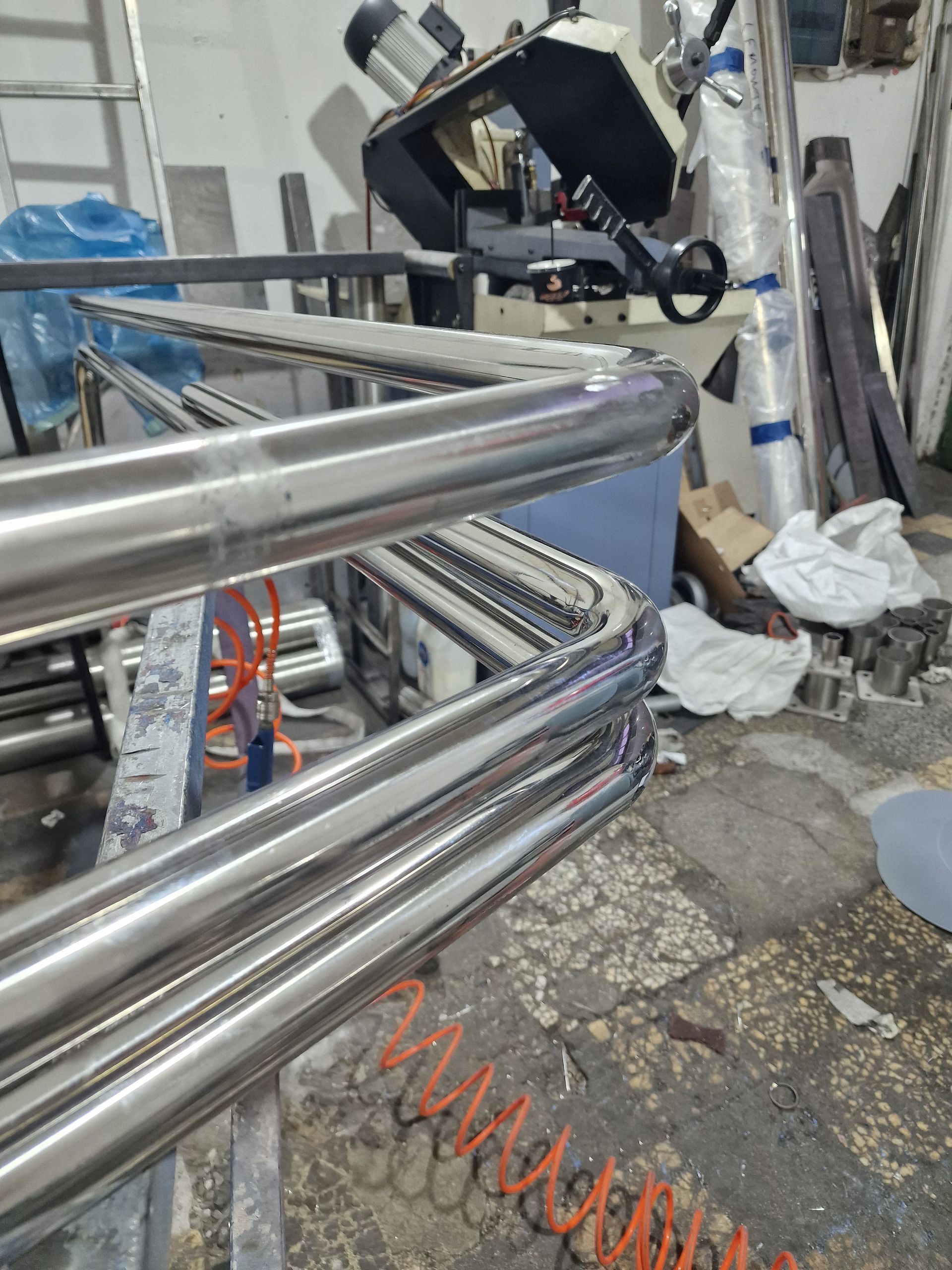 Καμπυλωτά, γυαλισμένα μεταλλικά εξαρτήματα κιγκλιδωμάτων στοιβαγμένα σε ένα εργαστήριο, με ένα μεταλλικό πριόνι στο βάθος.