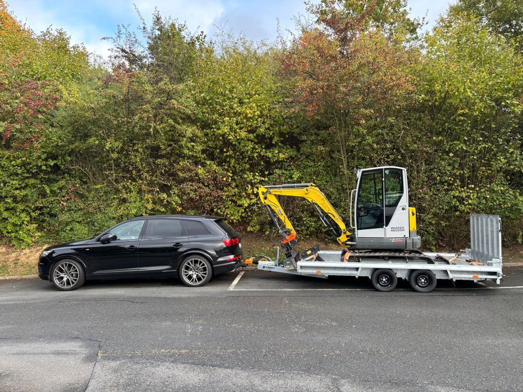 Ein schwarzes Auto zieht einen Anhänger mit einem kleinen gelben Bagger, der neben einer Reihe grüner Büsche parkt.
