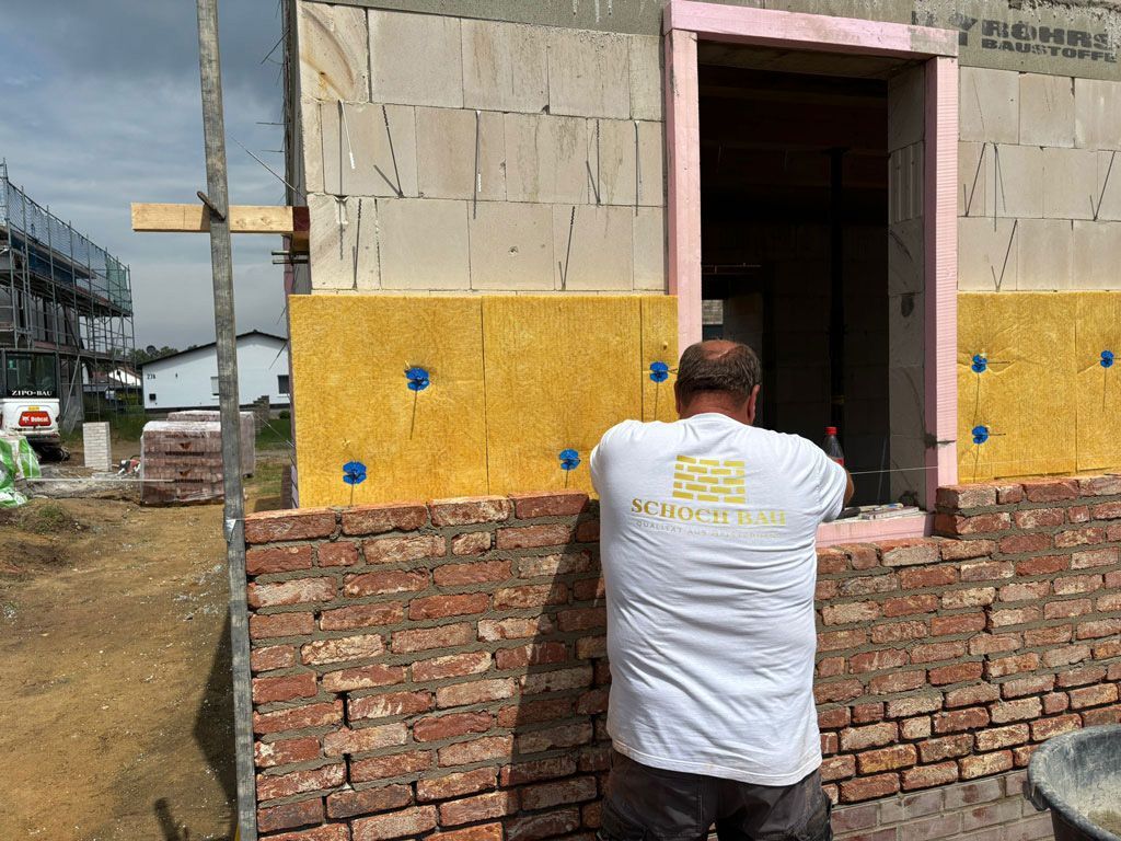 Ein Bauarbeiter bringt an der Außenfassade eines Gebäudes in der Nähe eines Fensters Dämmmaterial an. Mauerwerk und Gerüst sind sichtbar.