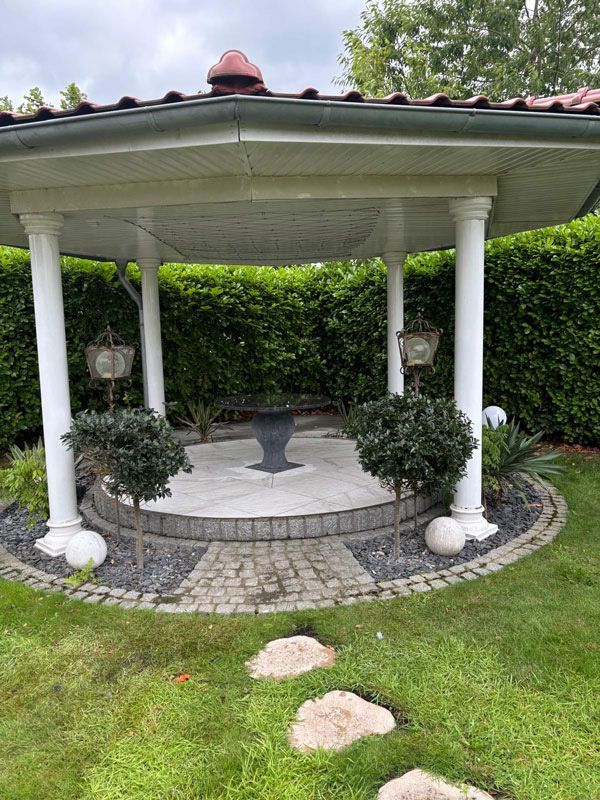 Pavillon mit weißen Säulen, rotbraunem Dach und gepflastertem Innenweg. Zwei kleine Bäume, ein gepflastertes Beet, eine grüne Hecke.