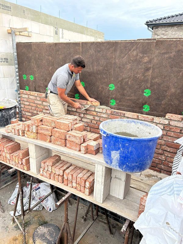 Eine Person verlegt Ziegelsteine ​​an einer Mauer; Ziegelsteine ​​und Mörtel liegen in der Nähe.