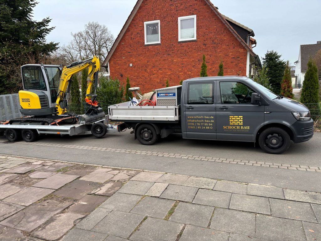 Ein grauer Lieferwagen zieht einen Anhänger mit einem kleinen Bagger auf einer Ziegelstraße vor einem Backsteinhaus.