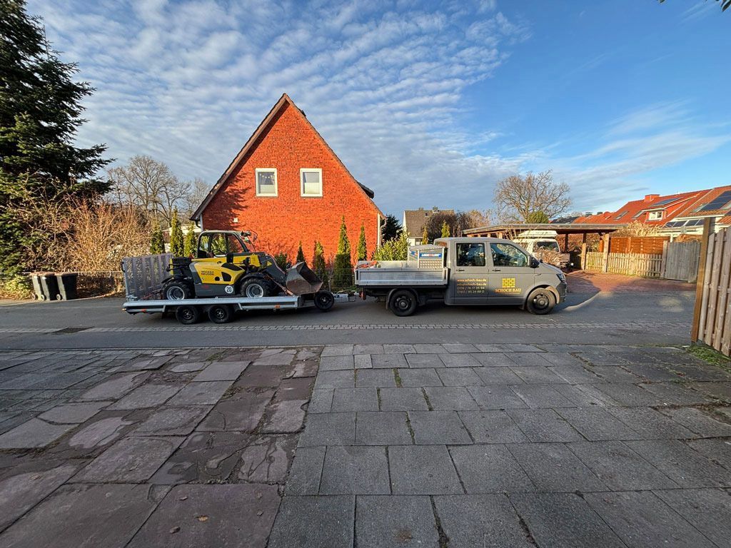 Ein Lastwagen mit Anhänger, auf dem ein gelber Bagger transportiert wird, parkt vor einem roten Backsteinhaus unter blauem Himmel.