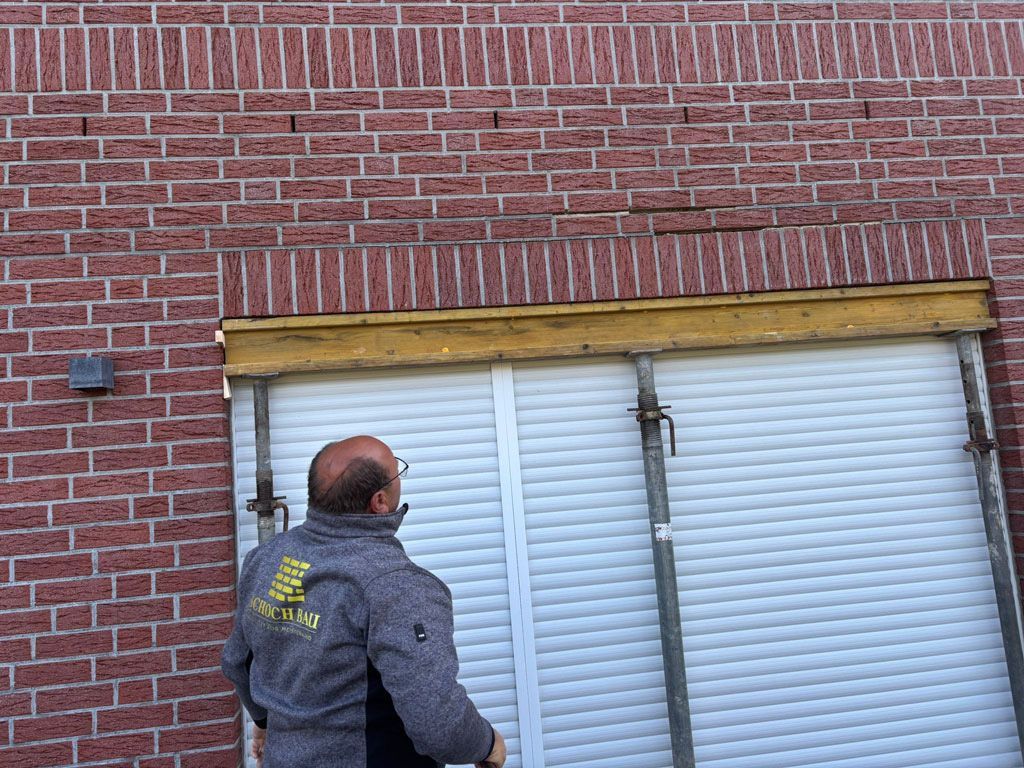 Ein Mann blickt zu einer Backsteinmauer mit weißem Fensterladen und Holzbalken hinauf.