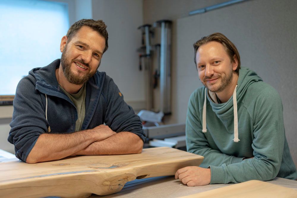 Zwei Männer sitzen mit verschränkten Armen an einem Holztisch.