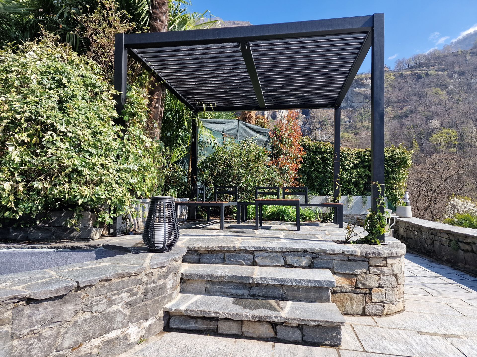 Pergolato su patio in pietra con posti a sedere, giardino, montagne sullo sfondo.