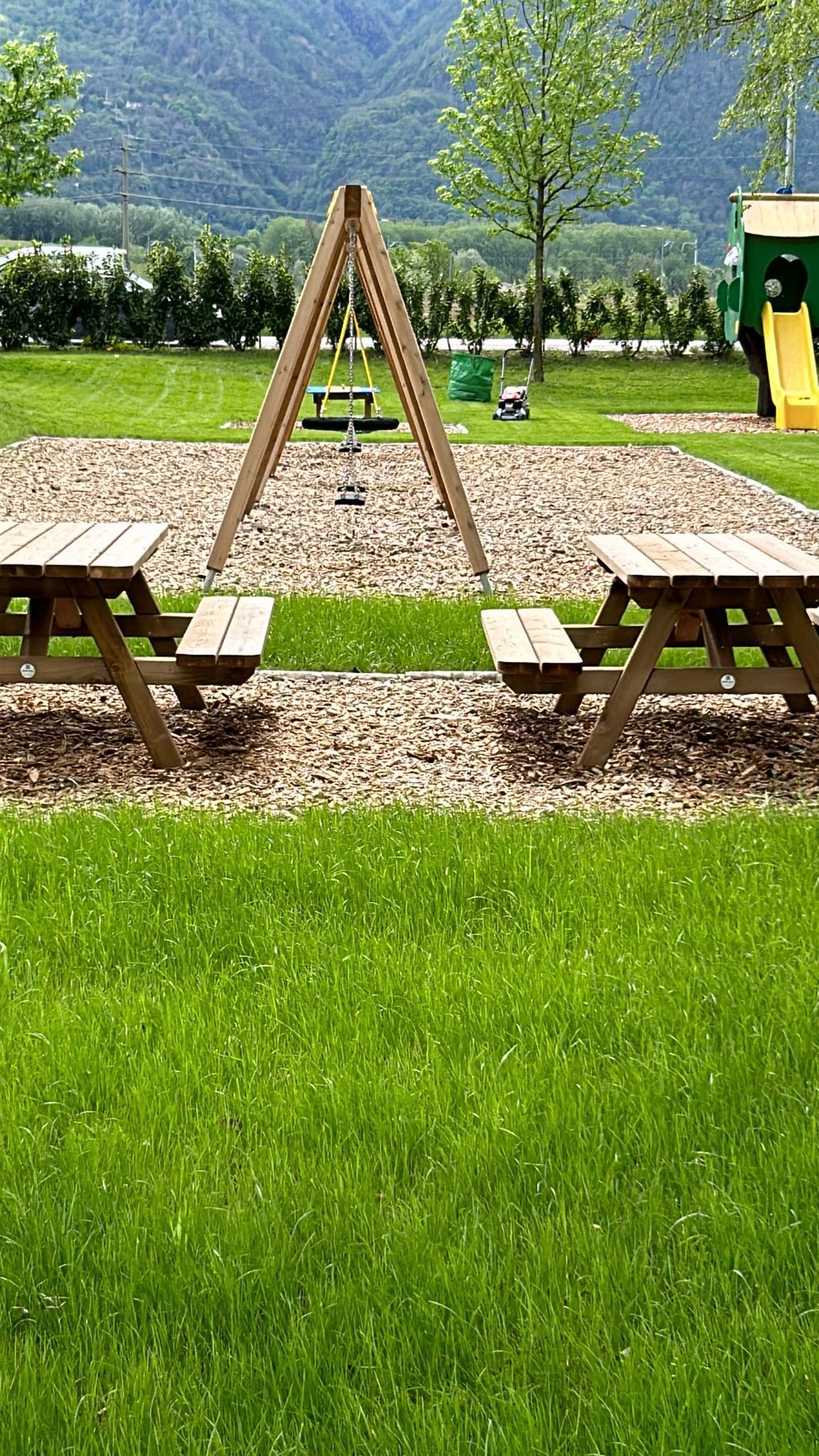 Area picnic all'aperto con tavoli in legno, altalena a forma di A, sentieri di ghiaia e prato verde in un contesto montano.