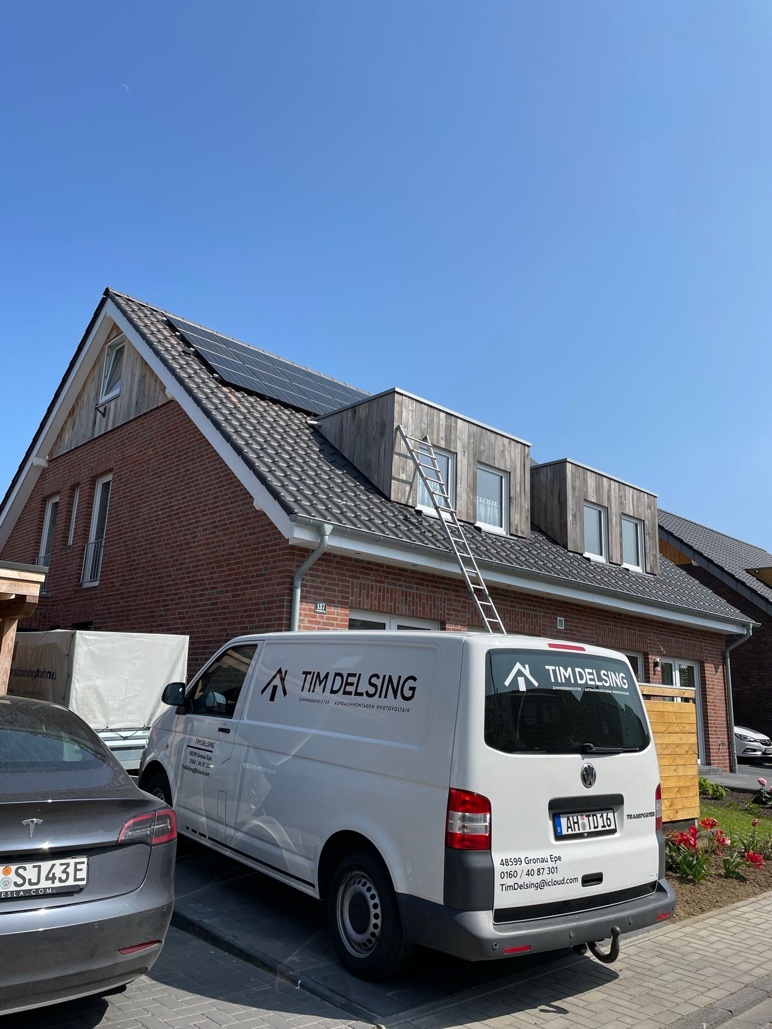 Ein weißer Lieferwagen parkt vor einem Backsteinhaus mit Solarzellen auf dem Dach.