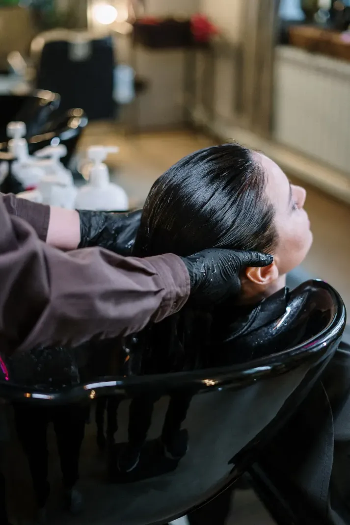 Una persona se lava el pelo en una peluquería. El estilista lleva guantes negros y una manga marrón; la persona en la silla parece relajada.