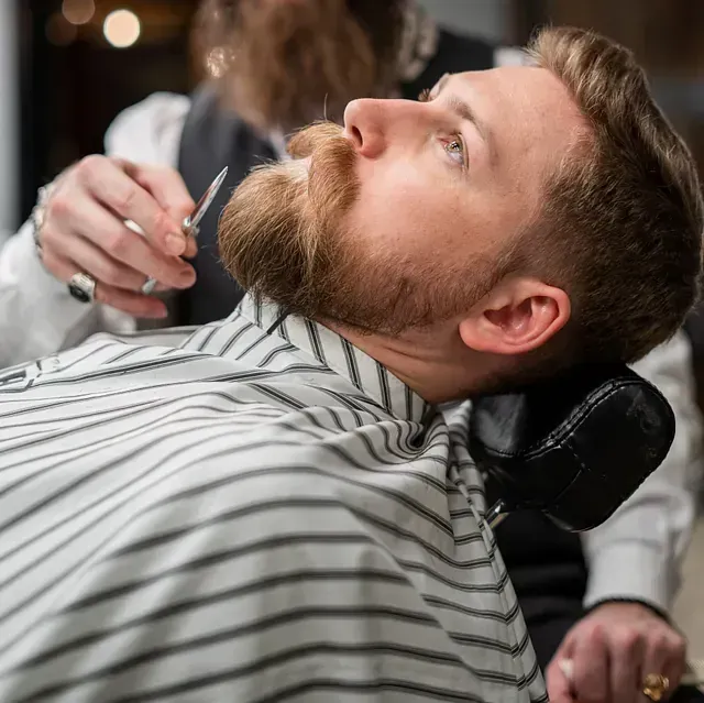 Un barbero le recorta la barba a un hombre. El hombre mira hacia arriba, con una capa de barbero a rayas.