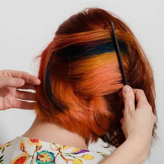 Persona peinándose el cabello con vibrantes reflejos naranjas y azules. Vista de espaldas, con las manos visibles, sobre fondo blanco.