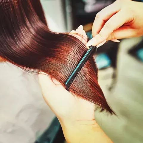 Unas manos peinan un cabello castaño rojizo brillante con un peine negro. El entorno parece ser interior, posiblemente un salón de belleza.
