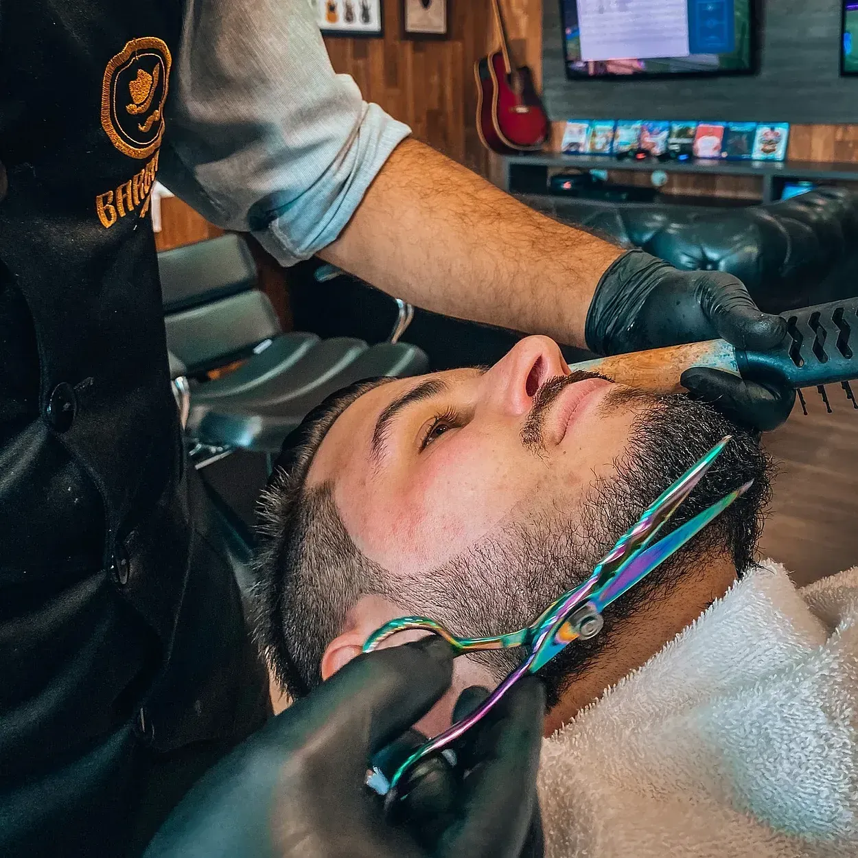 Un barbero recorta la barba de un hombre con tijeras en una barbería. El barbero lleva guantes y un delantal negro. El hombre está recostado, cubierto con una toalla y relajado.
