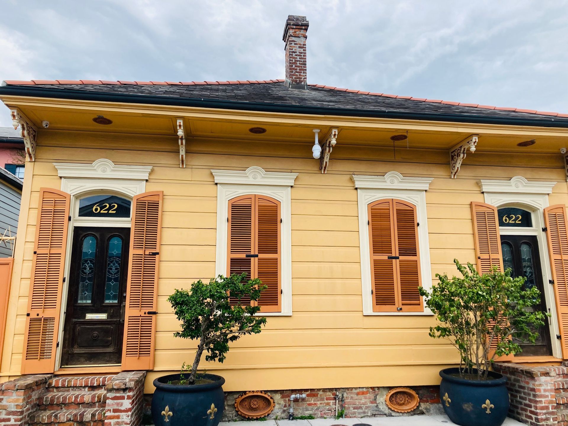 Yellow house facade with wooden shutters, blue doors, and potted plants by the entrance