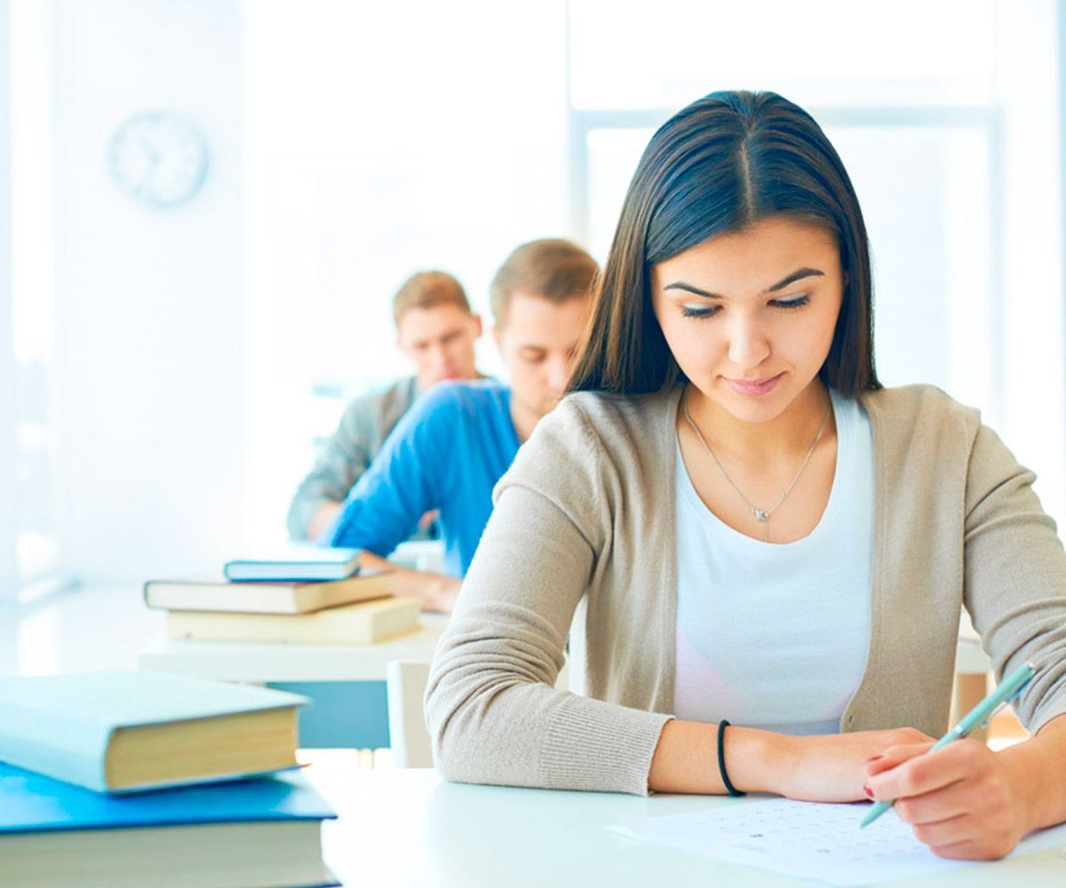 Una mujer está sentada en un escritorio en un aula escribiendo en un trozo de papel.