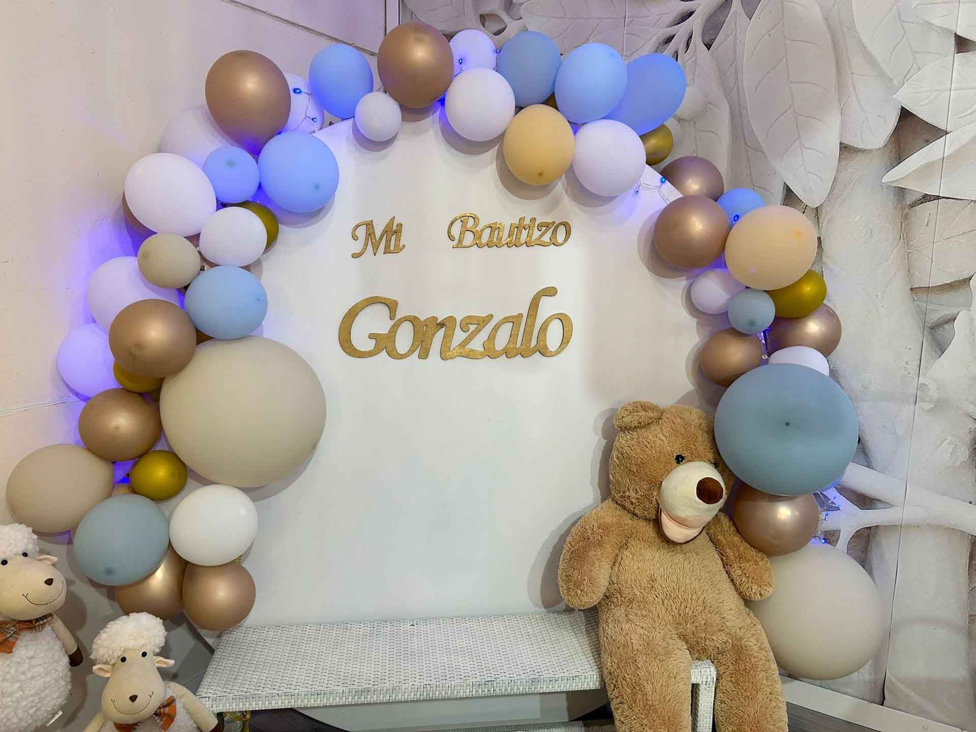 Un osito de peluche está sentado en un banco frente a una pared decorada con globos.