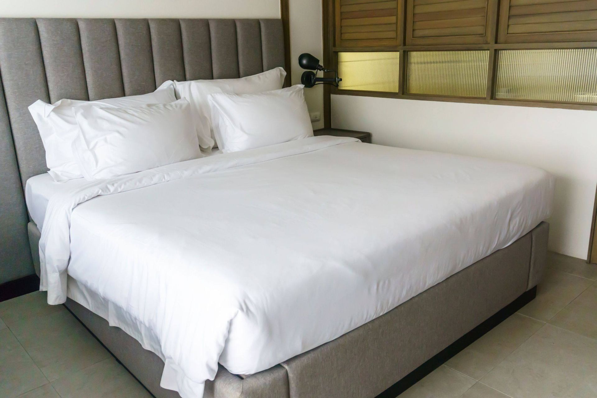 Una cama cuidadosamente hecha con sábanas y almohadas blancas; cabecera y estructura tapizadas en gris.