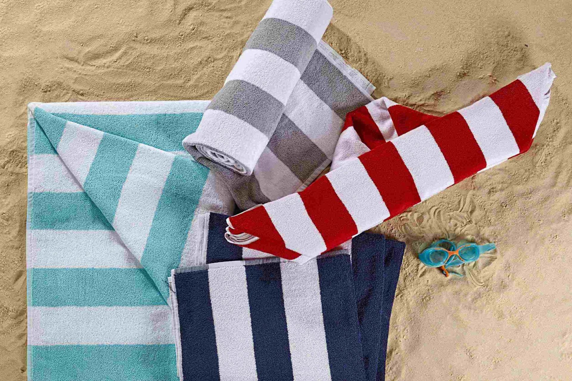 Toallas de playa en varios colores (azul, verde, rojo, gris) con rayas blancas sobre arena.