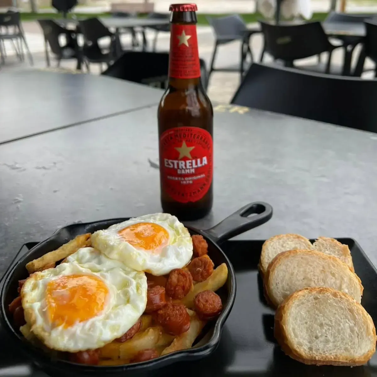Sartén de huevos fritos y salchichas sobre patatas, con cerveza y pan en una mesa.
