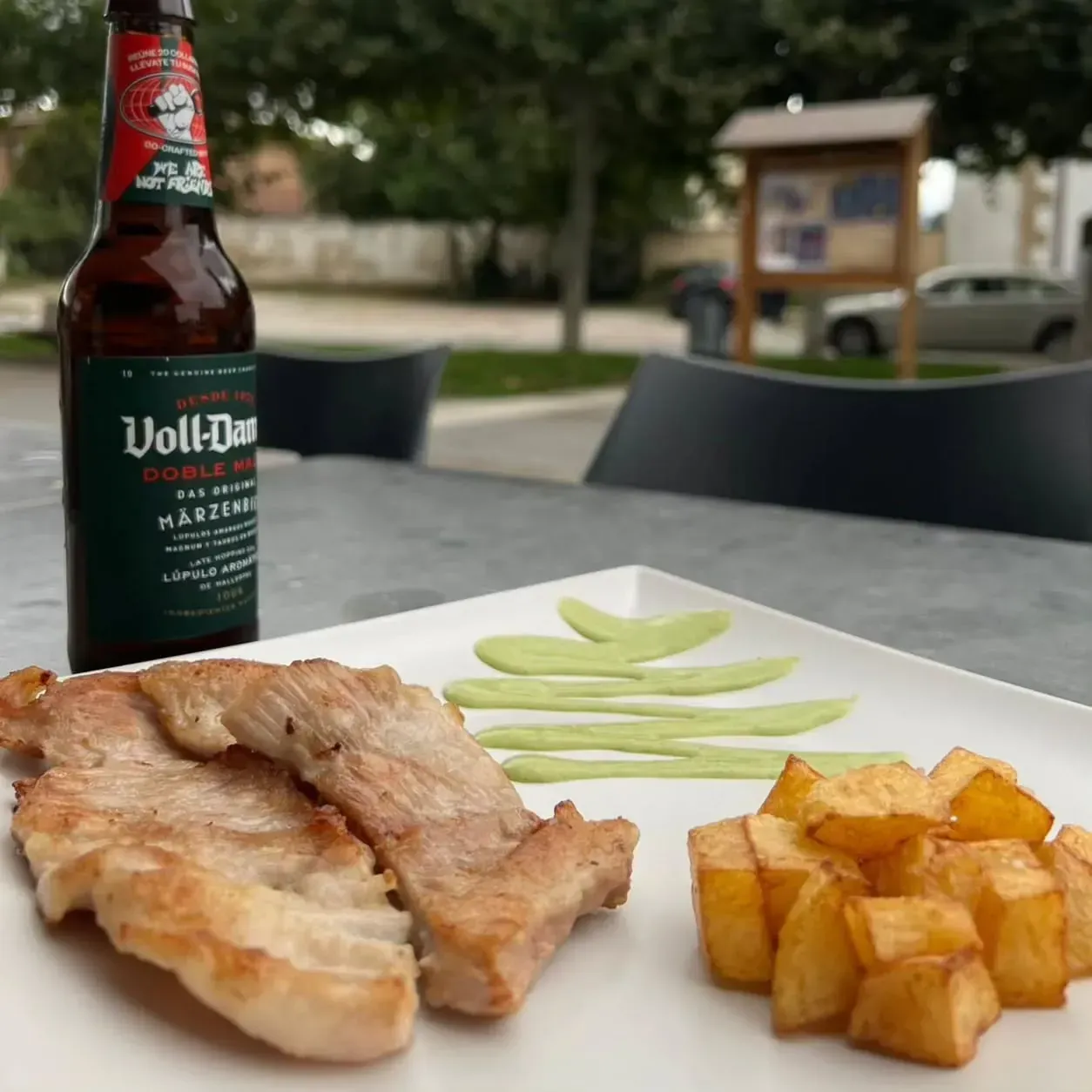 Plato de cerdo, patatas y salsa verde con botella de cerveza en la mesa al aire libre.